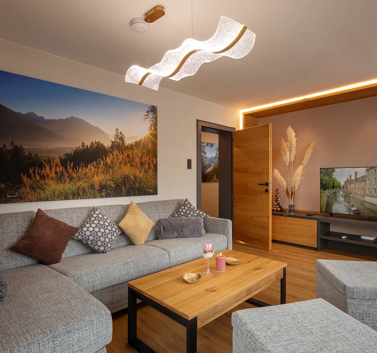 Modern living room with a gray sectional sofa, wooden coffee table, decorative pillows, a large landscape photo on the wall, and a TV on a wooden cabinet.