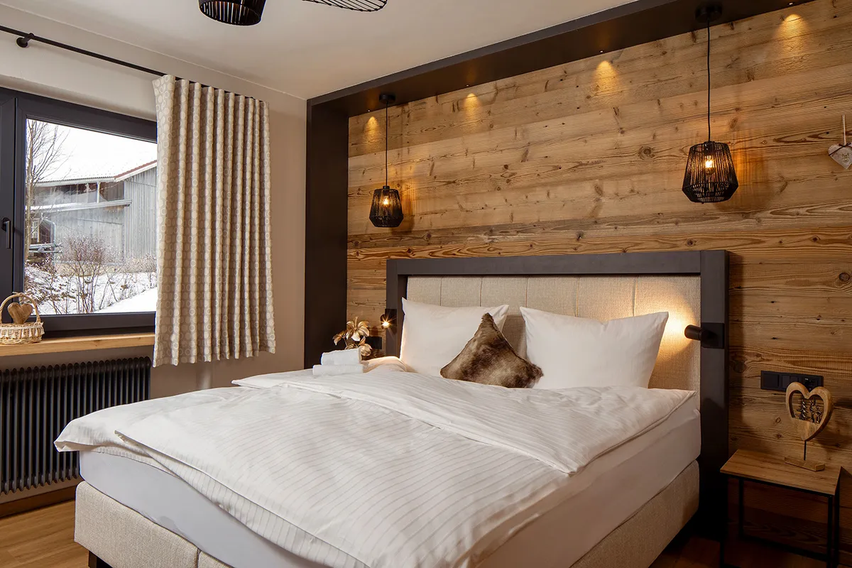 Cozy bedroom with a large bed featuring white linens, beige and brown pillows, wood-paneled accent wall, and pendant lights.
