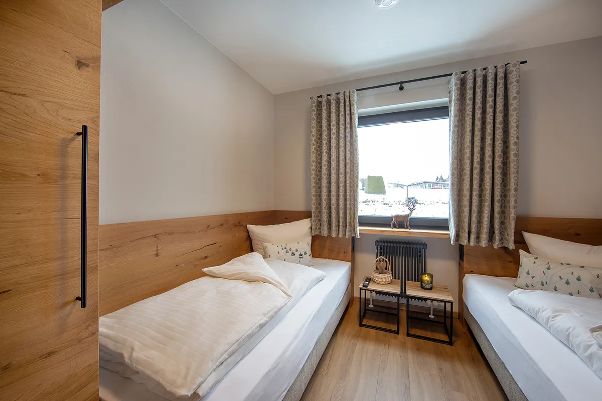 Small bedroom with two single beds, white bedding, wooden headboards, a window with patterned curtains, and a small table with a candle, basket, and remote control.