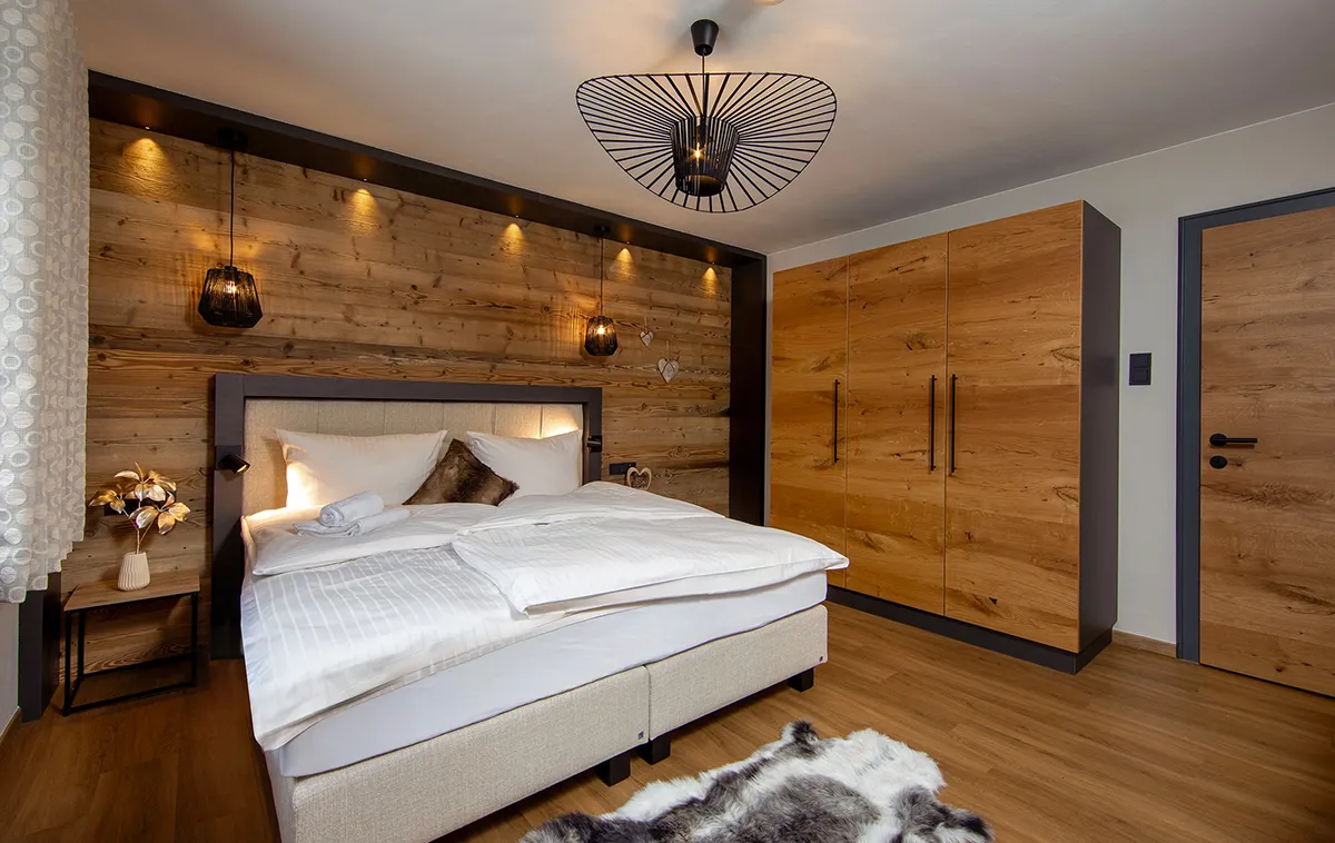 Cozy bedroom with wooden walls, beige bed with white linens, fur pillow, black pendant lights, wooden wardrobe, and furry rug on wooden floor.