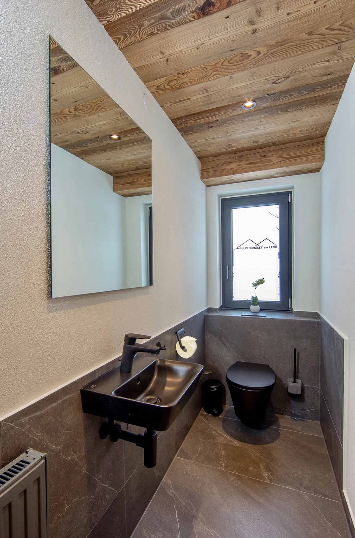 Modern small bathroom with black wall-mounted sink, black toilet, gray tiles, wooden ceiling, and a window with a small plant on the sill.