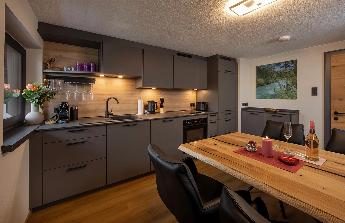 Modern kitchen with gray cabinets, wooden backsplash, black countertop appliances, and a wooden dining table set with candles, wine glasses, and a bottle.