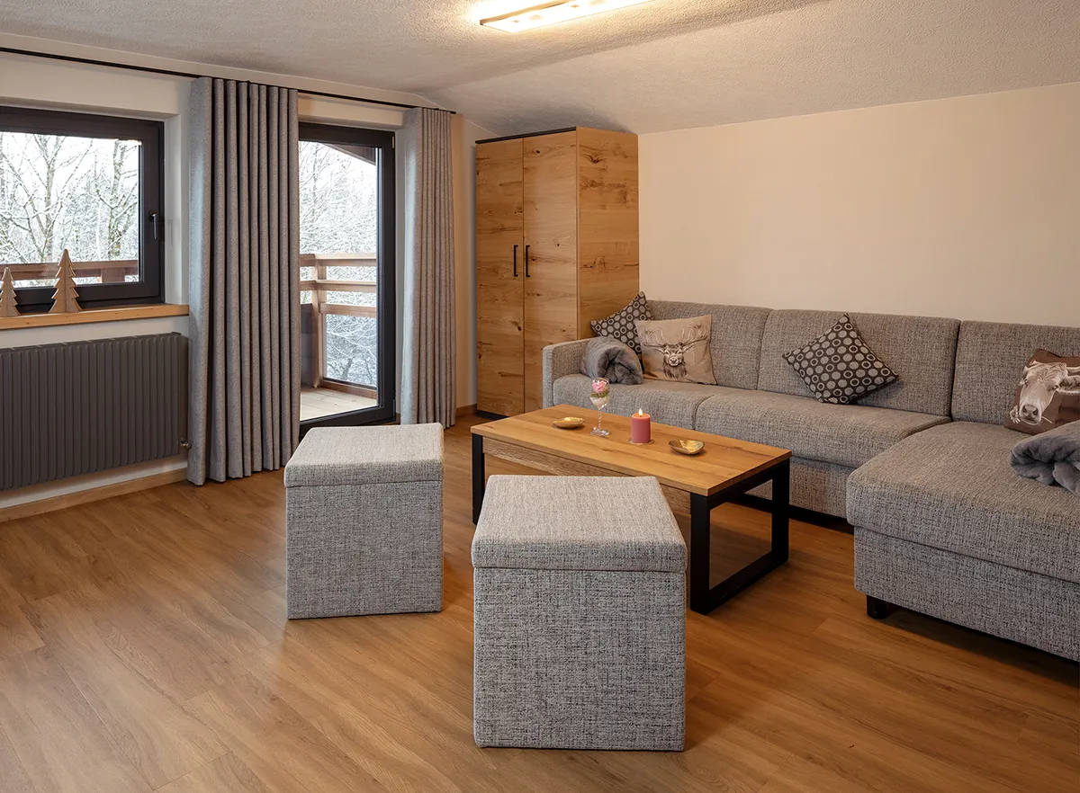 Cozy living room with gray sectional sofa, two gray ottomans, wooden coffee table, wooden cabinet, and a window showing snowy trees outside.