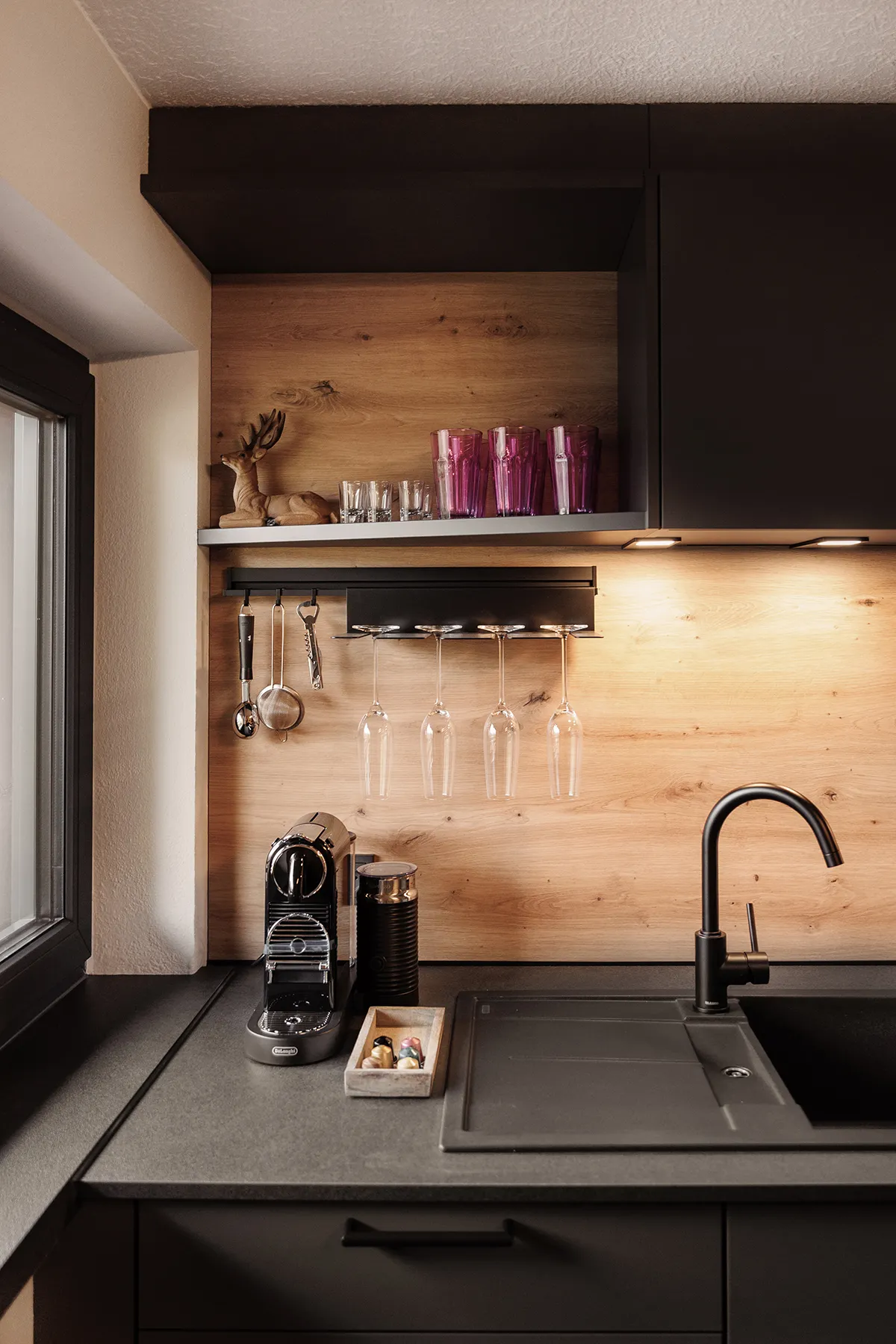 Modern kitchen corner with black countertop, black faucet, coffee machine, hanging wine glasses, and wooden shelf with decorative deer and pink glasses.