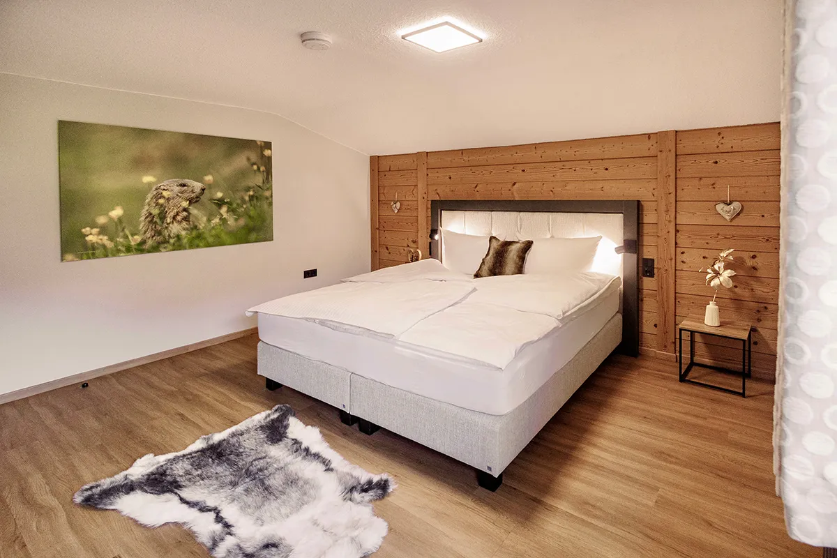 Cozy bedroom with wooden floor, double bed with white linens, fur pillow, animal print rug, and nature wall art.