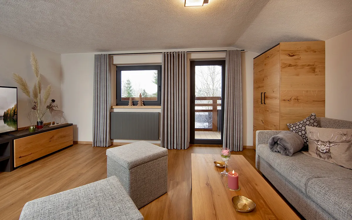 Cozy living room with grey upholstered sofa, wooden coffee table with a pink candle and flower, ottomans, wooden cabinet, window with grey curtains, and TV on a wooden stand.
