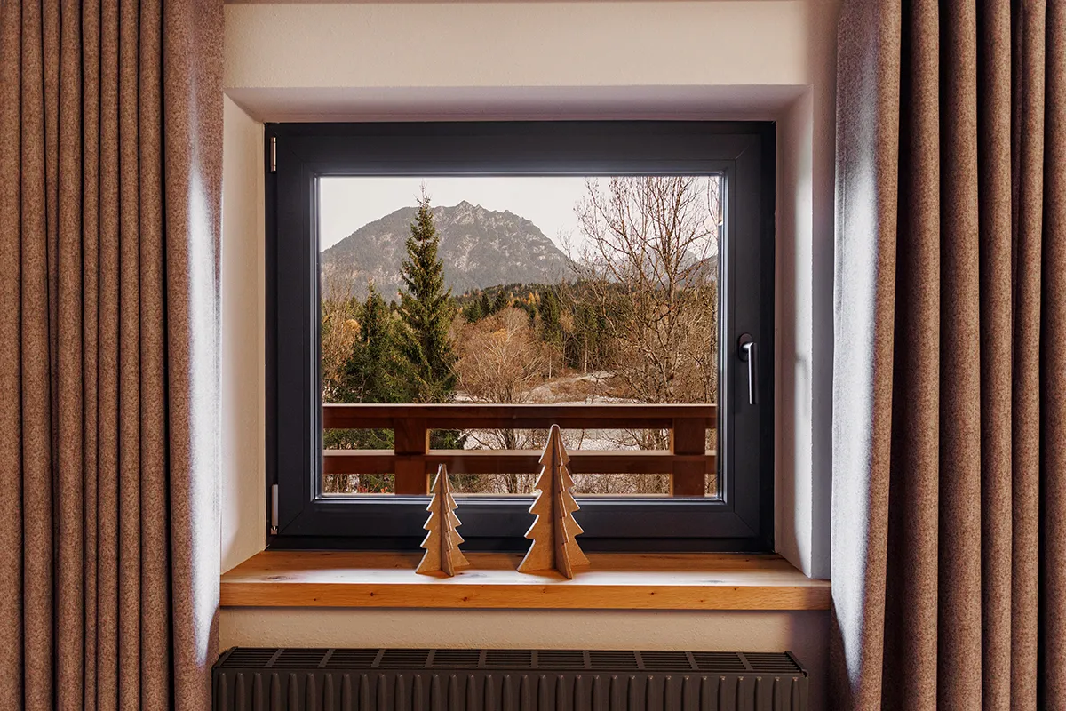 View through a black-framed window showing a mountain and forest, with two wooden decorative trees on the windowsill and brown curtains on each side.