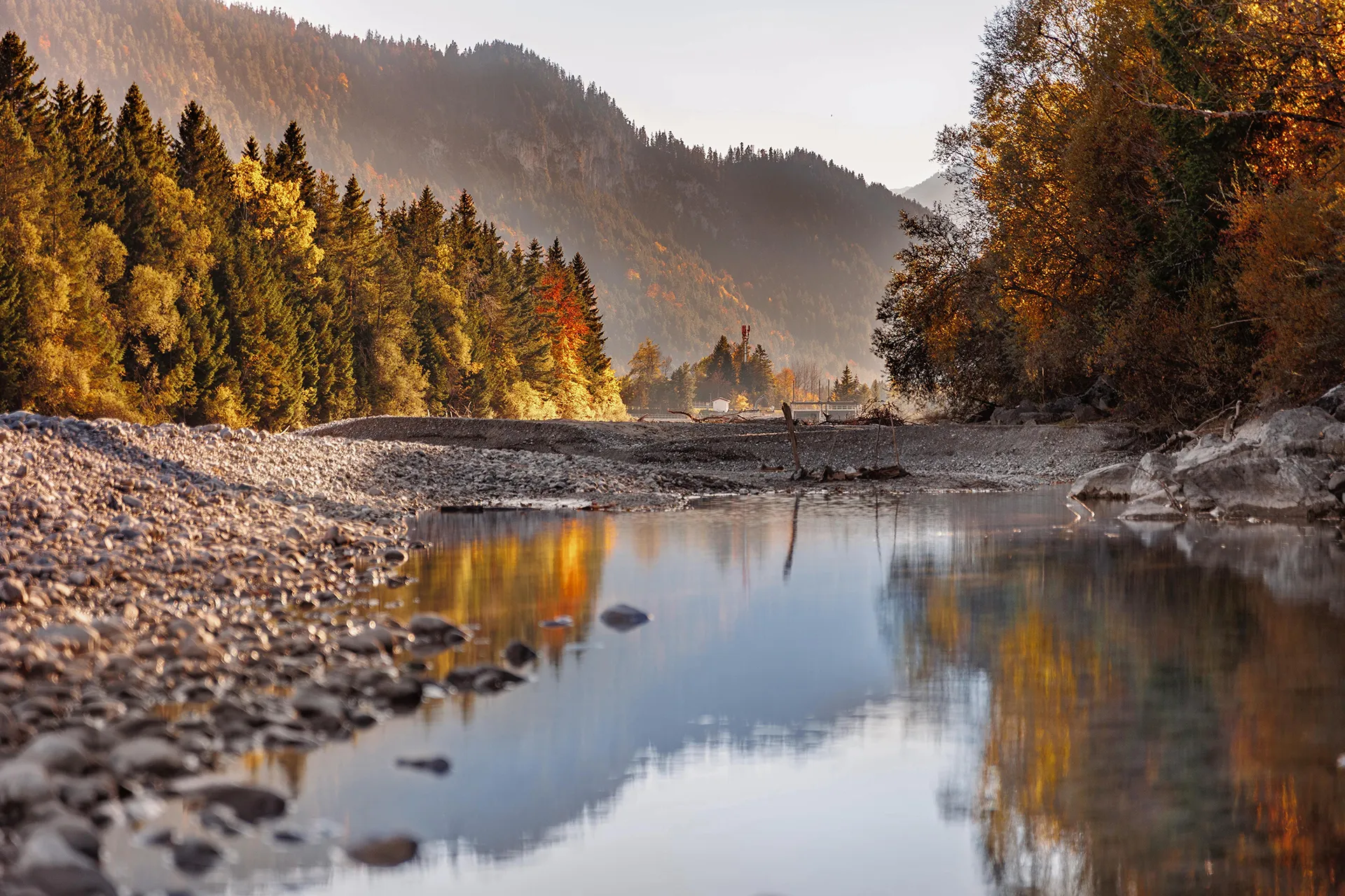 Calm river reflecting autumn trees and mountains under soft sunlight.