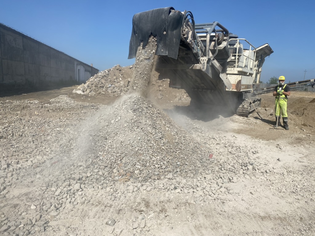 Heavy machinery dumping crushed rocks on ground with worker in yellow safety gear nearby.