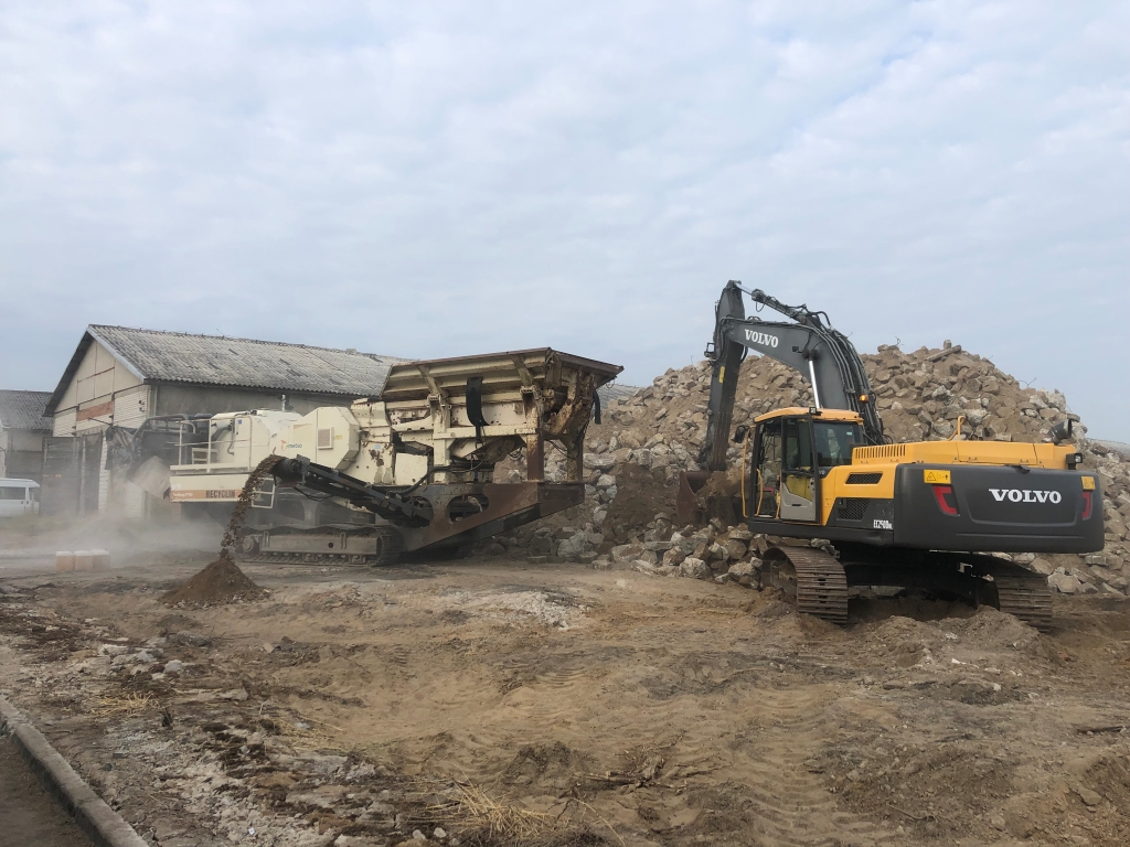 Yellow Volvo excavator lifting rocks near a machine sorting rubble at a demolition site with a cloudy sky.