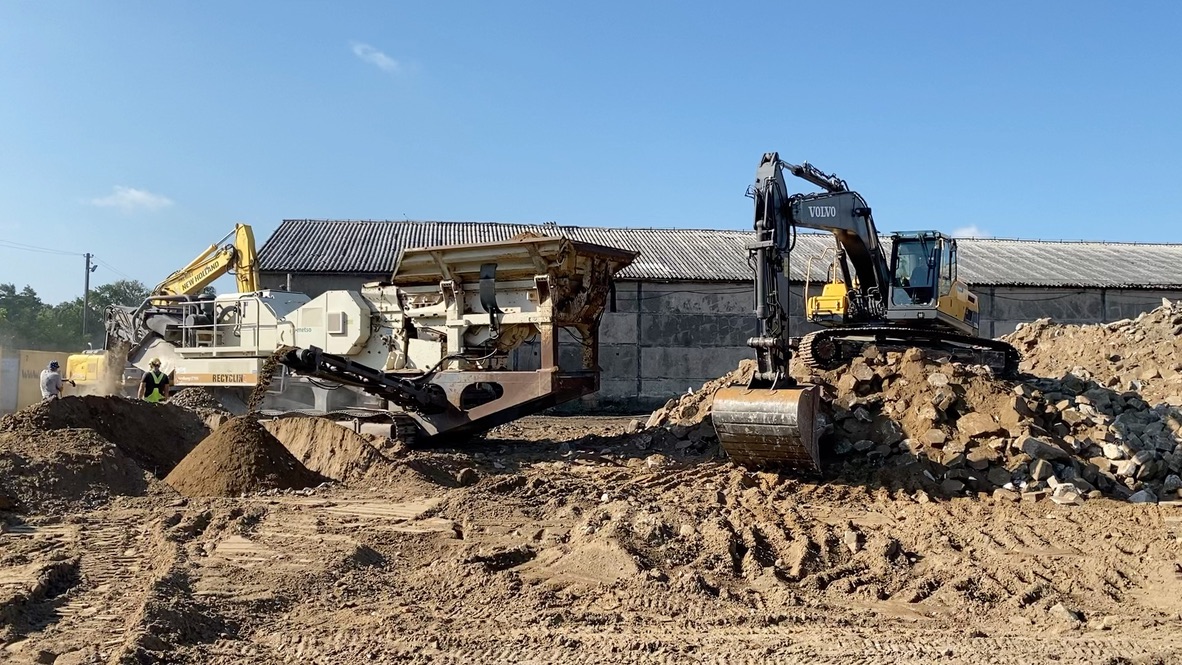 Excavator and rock crusher working on piles of dirt and rocks at a construction site under clear sky.