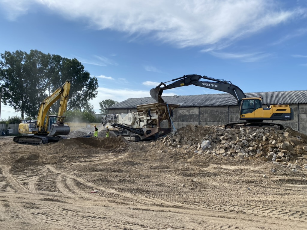 Two excavators working on a construction site with piles of rocks and two workers in safety gear.