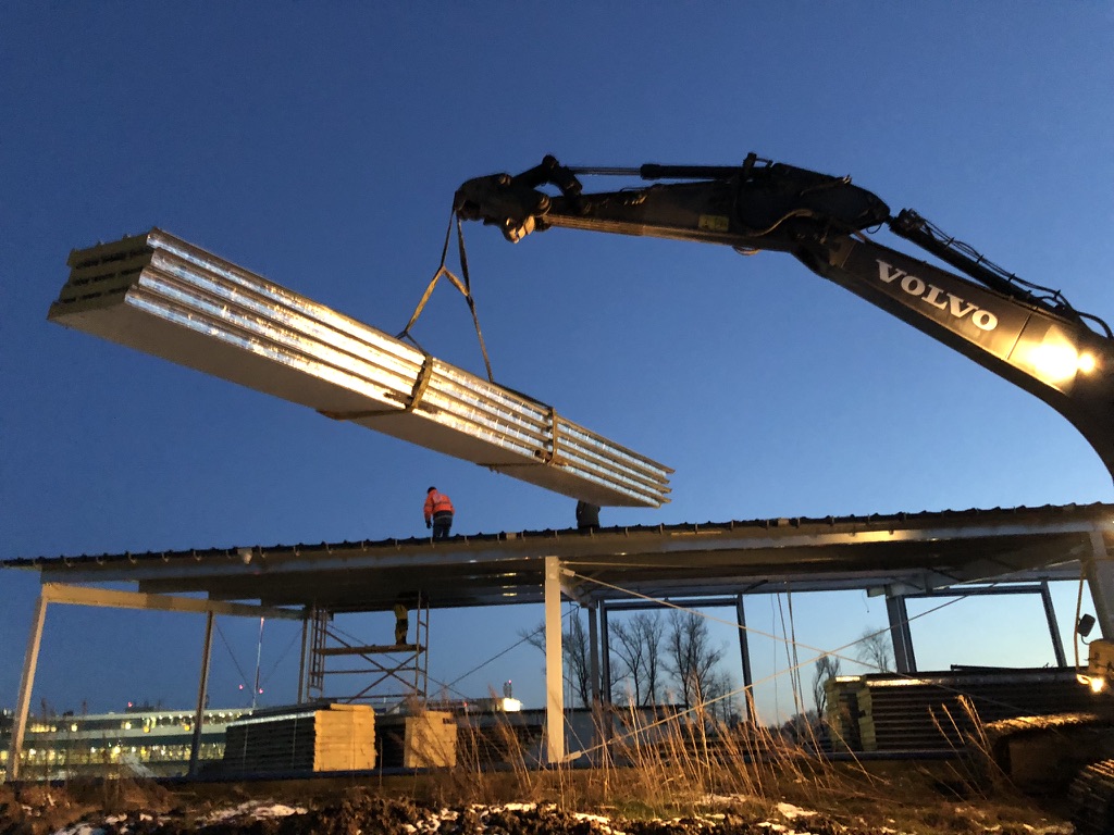 Excavator lifts metal beams while workers guide them onto a steel structure at dusk.