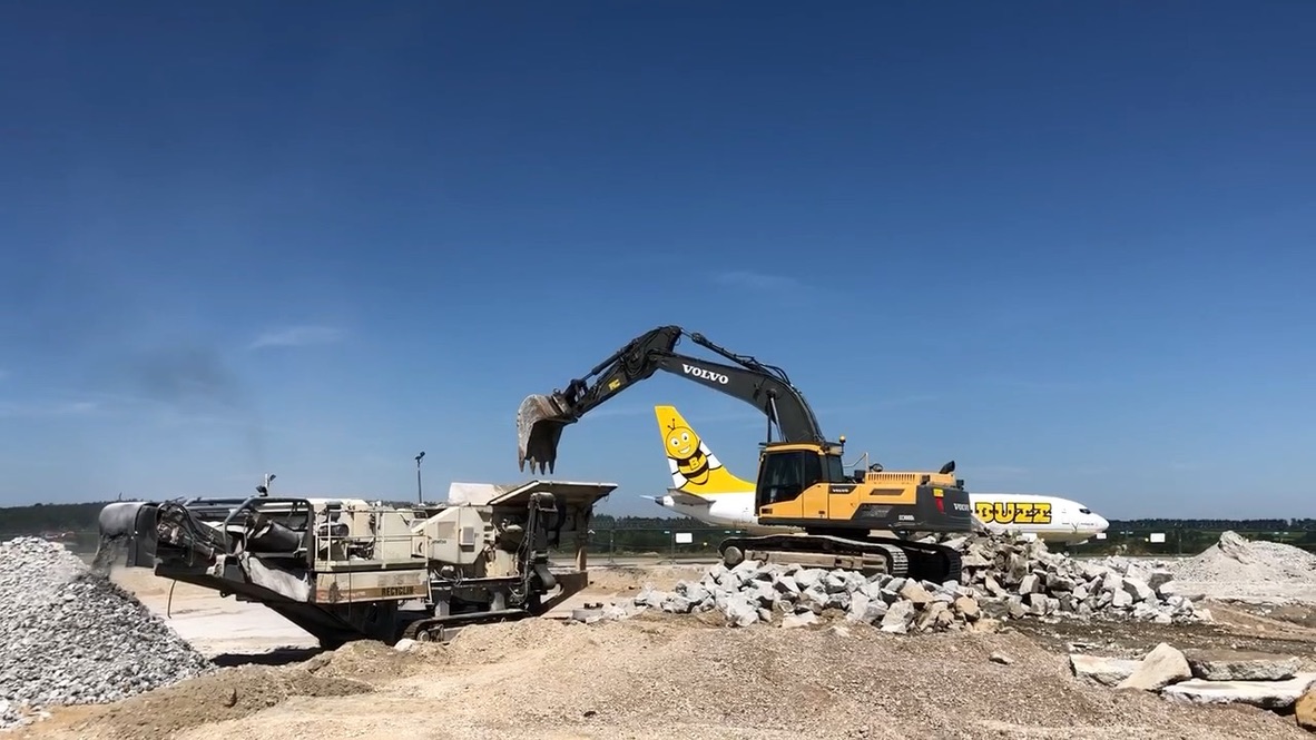 Yellow Volvo excavator loads rubble near a white plane with a smiling bee tail logo on a clear day.