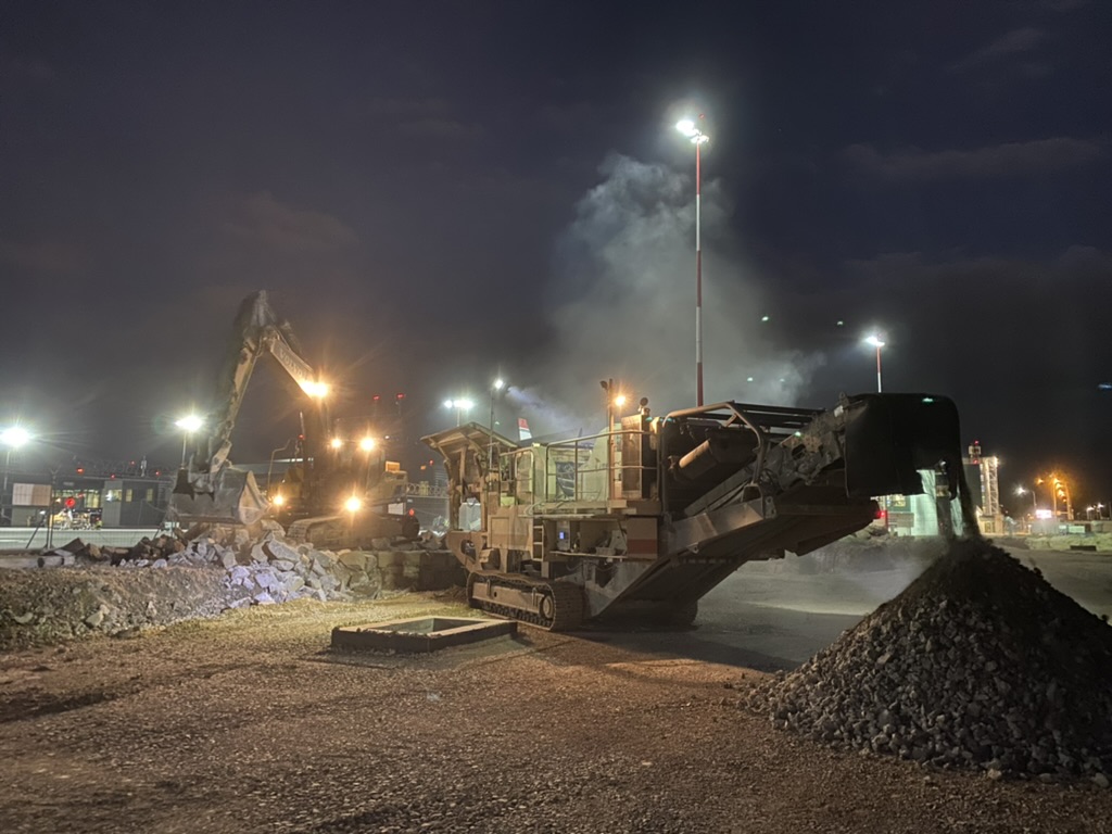 Construction site at night with excavator and conveyor machine piling gravel under bright lights.