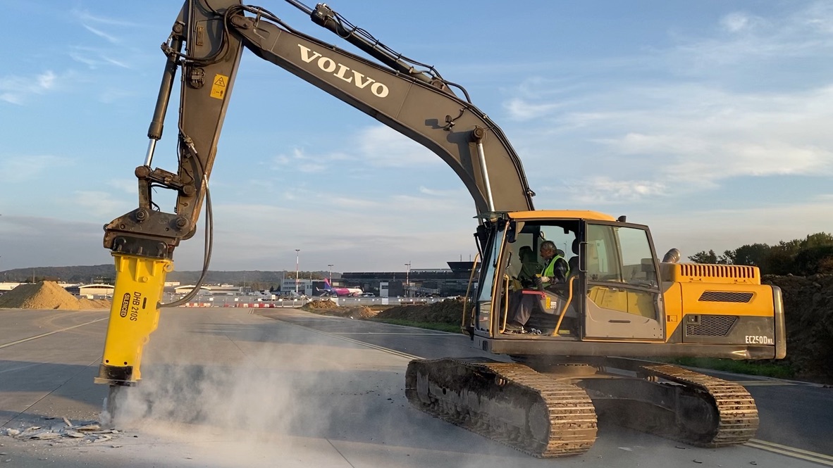Volvo excavator with hydraulic breaker demolishing concrete on a construction site with operator inside cabin.