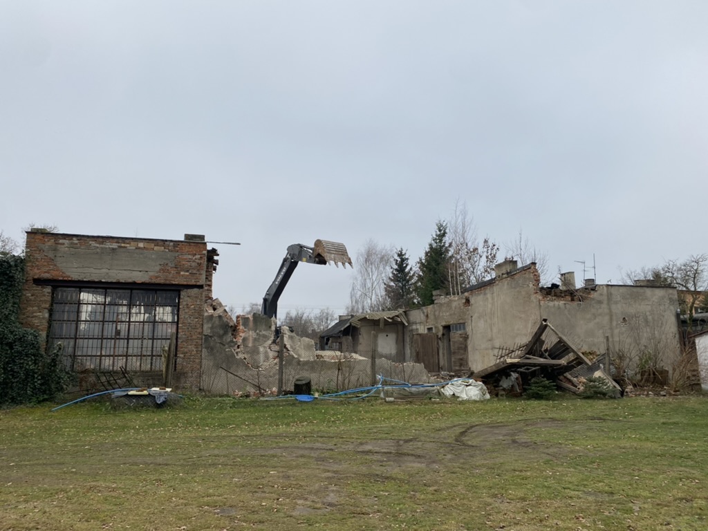 Partially demolished brick and concrete building with excavator arm lifting debris on overcast day.