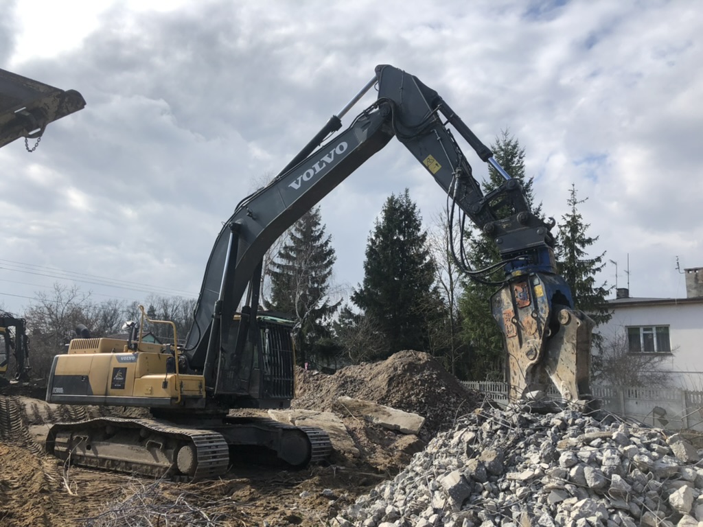 Volvo excavator with hydraulic crusher attachment breaking rubble at construction site with trees and a house.