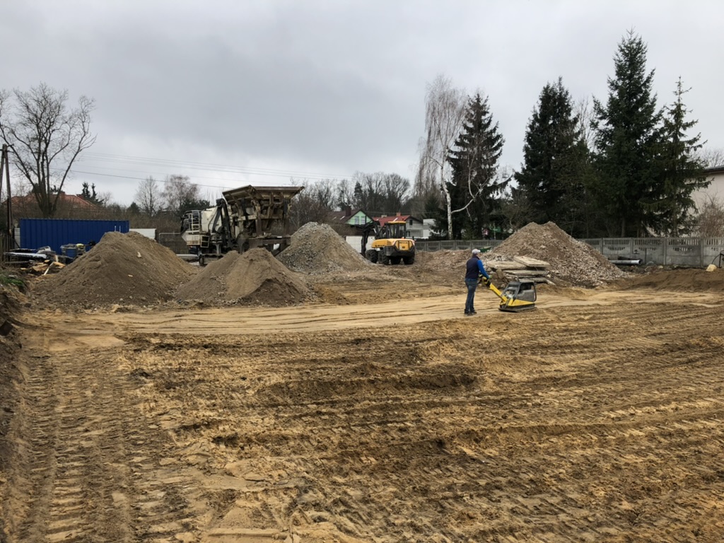 Construction site with piles of dirt, machinery, and a worker operating a compactor on muddy ground.
