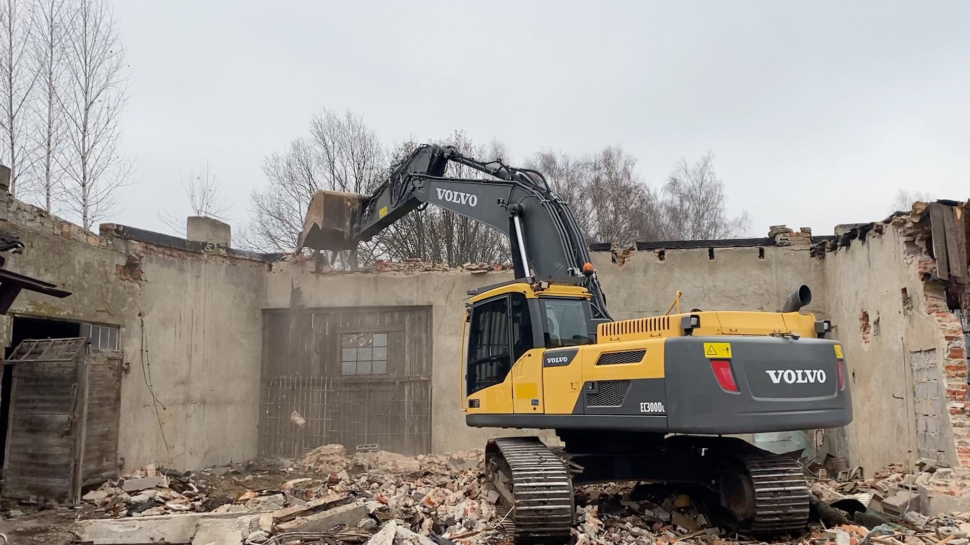Yellow Volvo excavator demolishing a brick building with rubble scattered around.