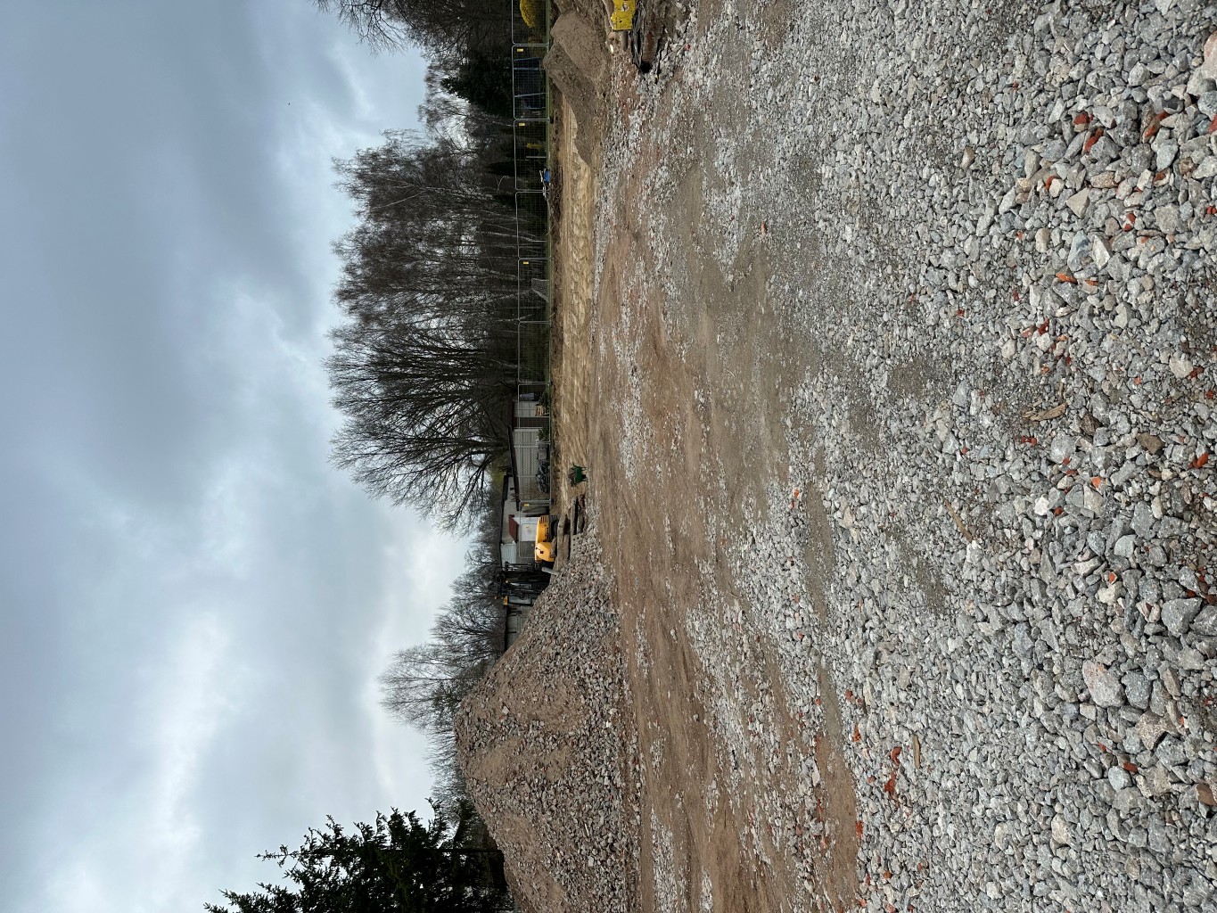 Construction site with gravel piles, a yellow excavator, and bare trees under a cloudy sky.