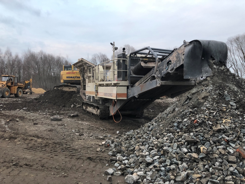 Construction site with machinery processing and piling crushed rocks under a cloudy sky.