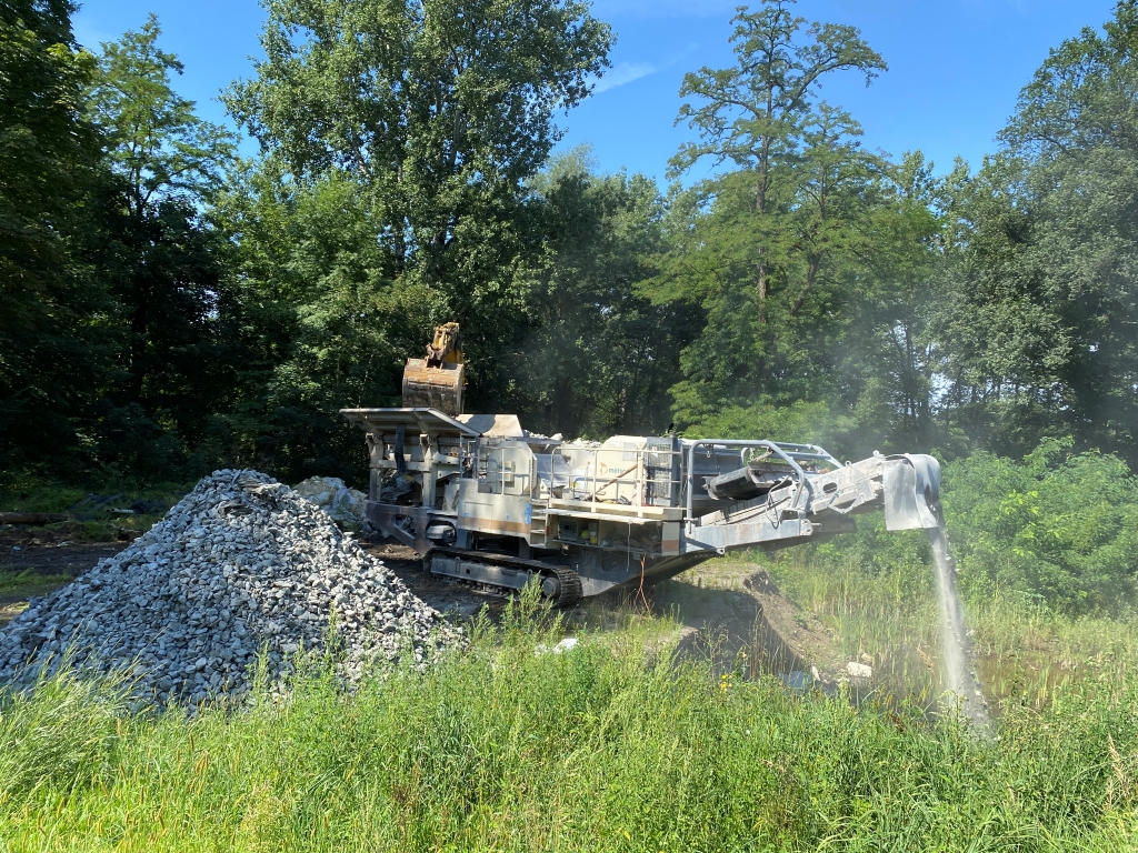 Machine crushing rocks with pile of crushed stones beside it in a green area under blue sky.