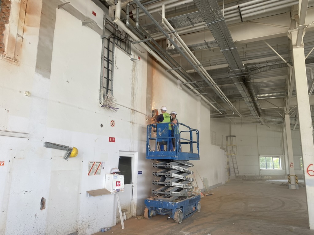 Two construction workers in safety gear on a blue scissor lift welding a wall inside a large industrial space.