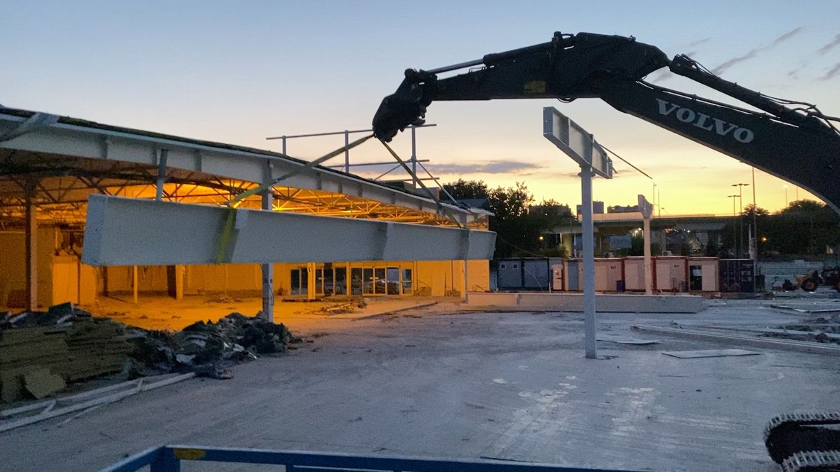Construction site at dusk with a Volvo excavator lifting a large steel beam near a partially built structure.