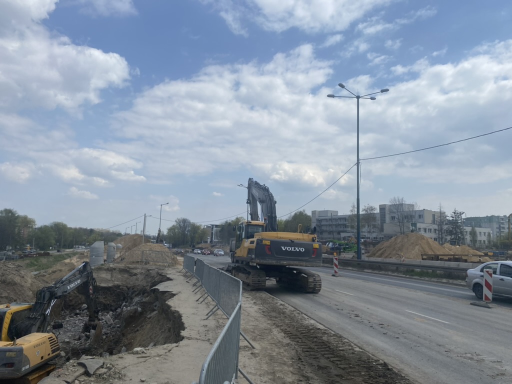 Construction site with excavators and dirt piles on a road under partly cloudy blue sky.