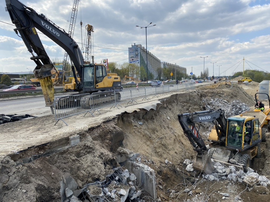Excavators demolishing concrete and digging earth at a roadside construction site on a cloudy day.