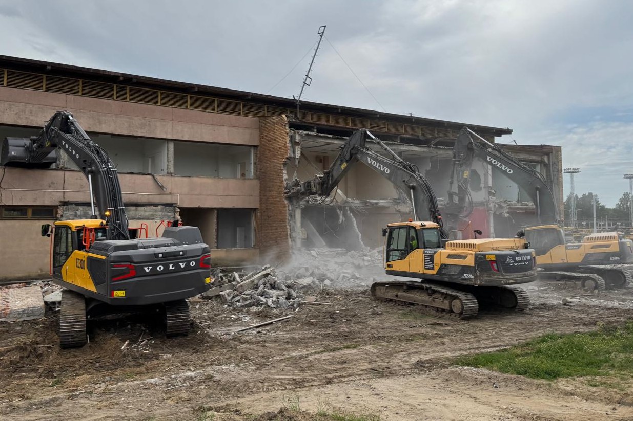 Three yellow Volvo excavators demolishing a large building with debris and dust around.