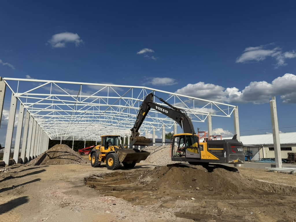 Construction site with Volvo excavator and loader working under white steel framework on dirt ground.