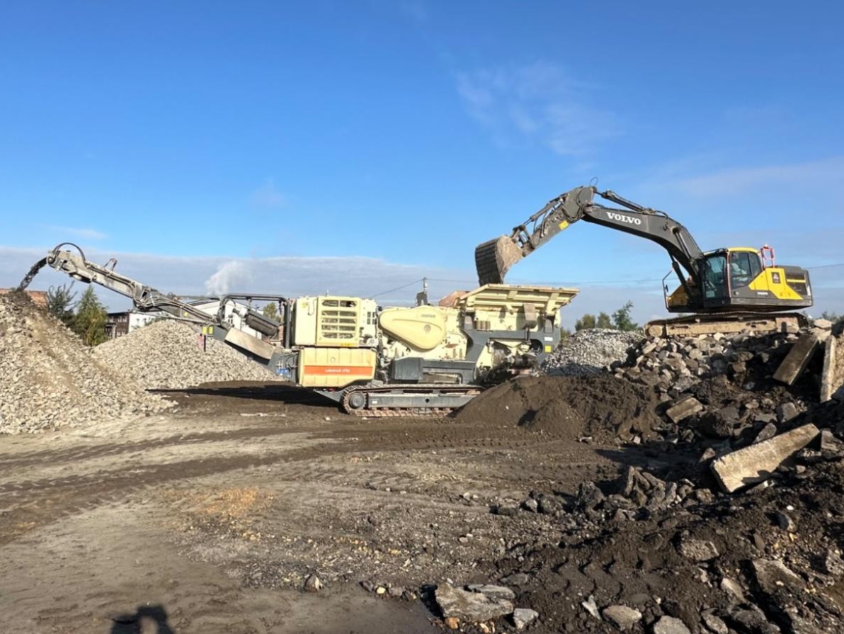 Excavator loading dirt into a crusher machine at a construction site with piles of gravel and dirt.