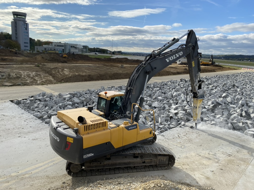 Yellow Volvo excavator with hydraulic tool working on concrete near large rocks at a construction site.