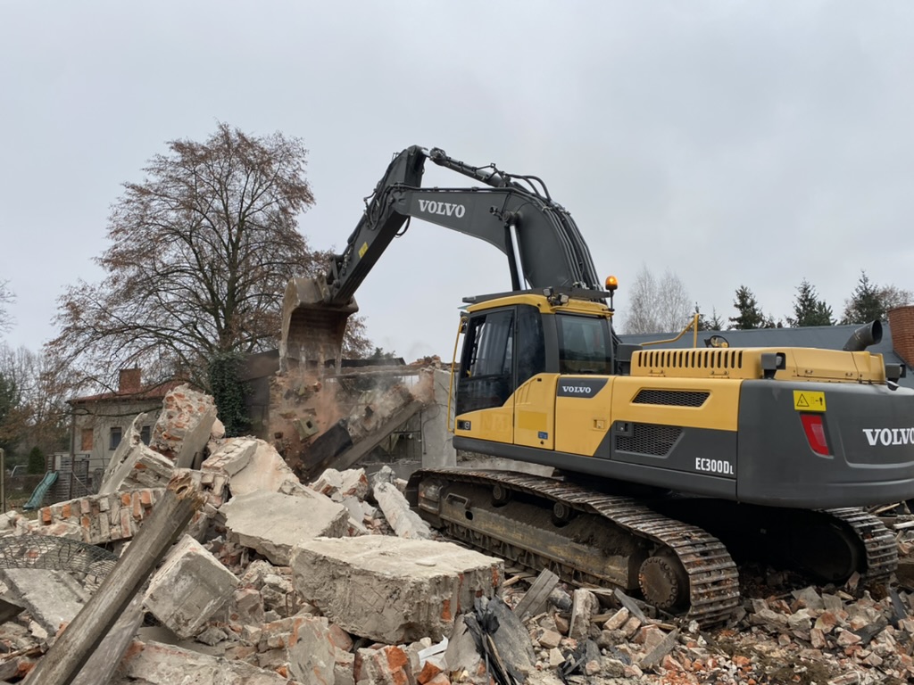 Yellow Volvo excavator demolishing a brick and concrete structure amid rubble under cloudy sky.