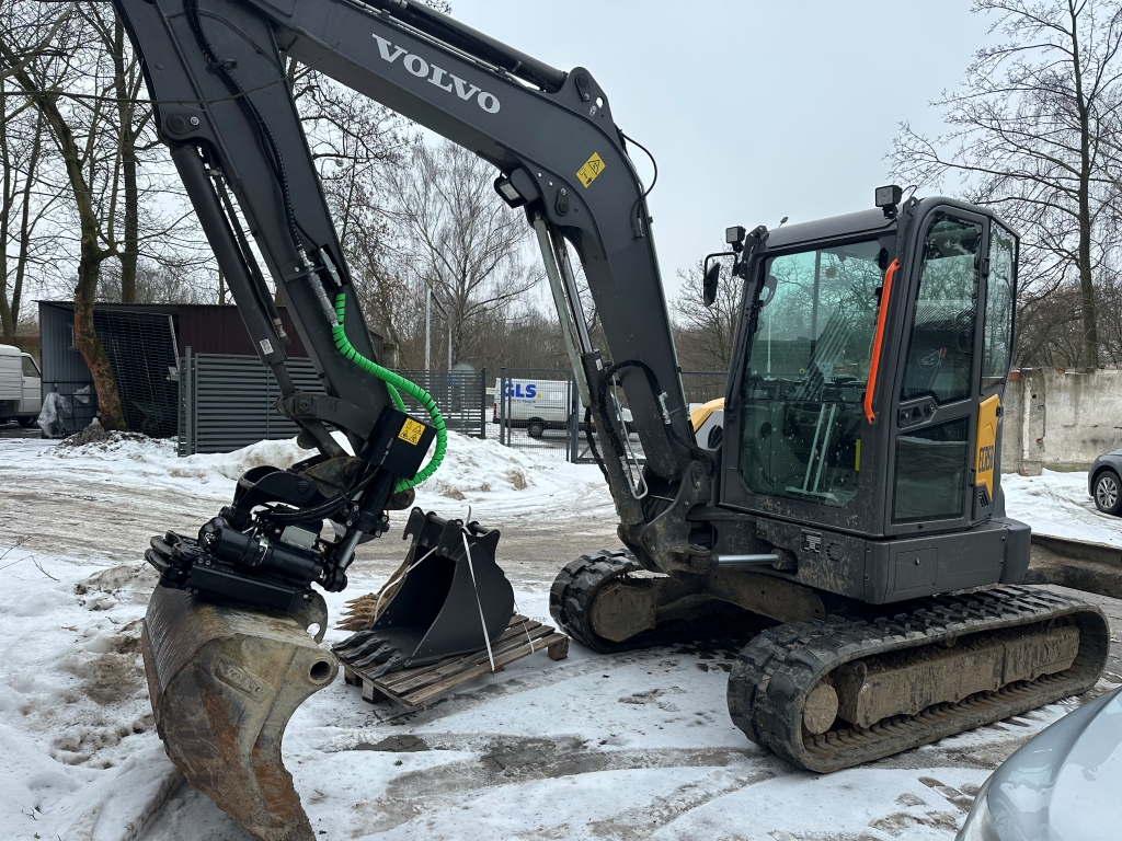 Volvo mini excavator parked on snow with extra bucket attachments on a pallet nearby.