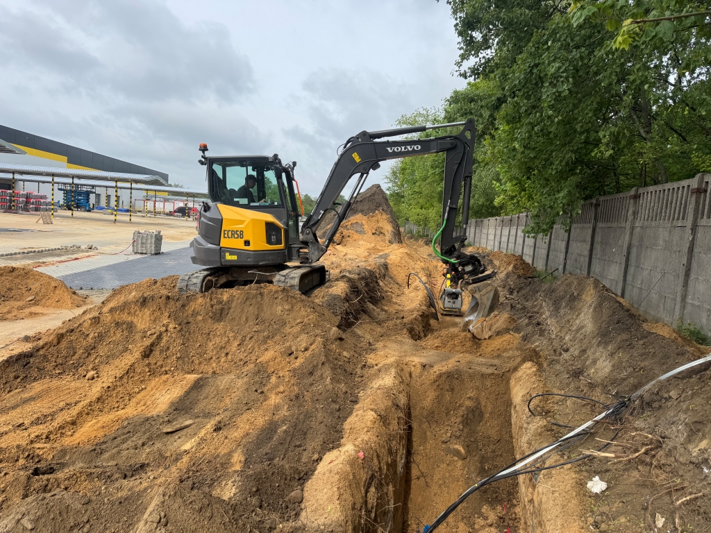 Yellow Volvo excavator digging a deep trench at a construction site near a concrete fence and trees.