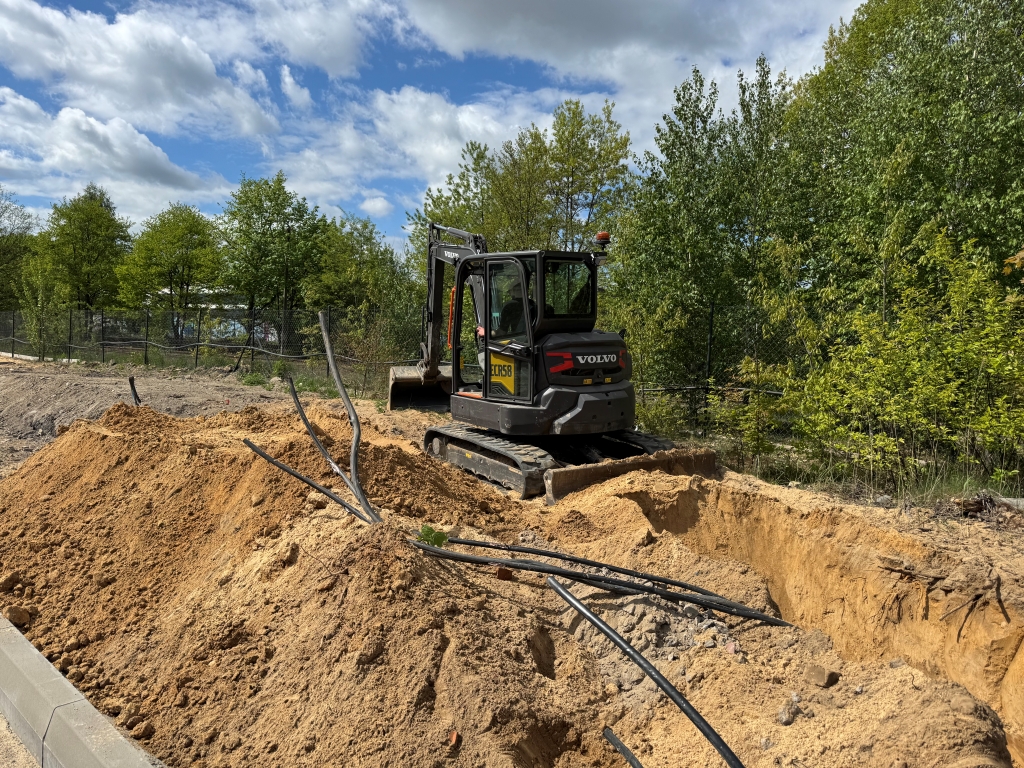 Black Volvo excavator on a construction site digging dirt with scattered black pipes around it.