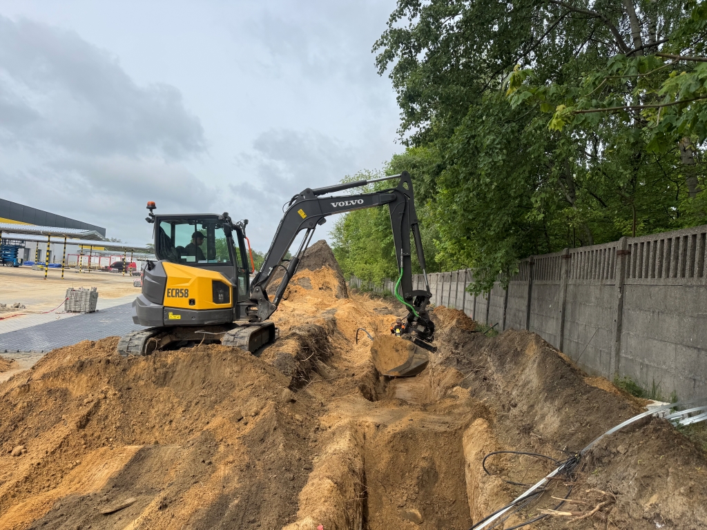 Yellow Volvo ECR58 excavator digging a trench beside a concrete fence with trees in background.