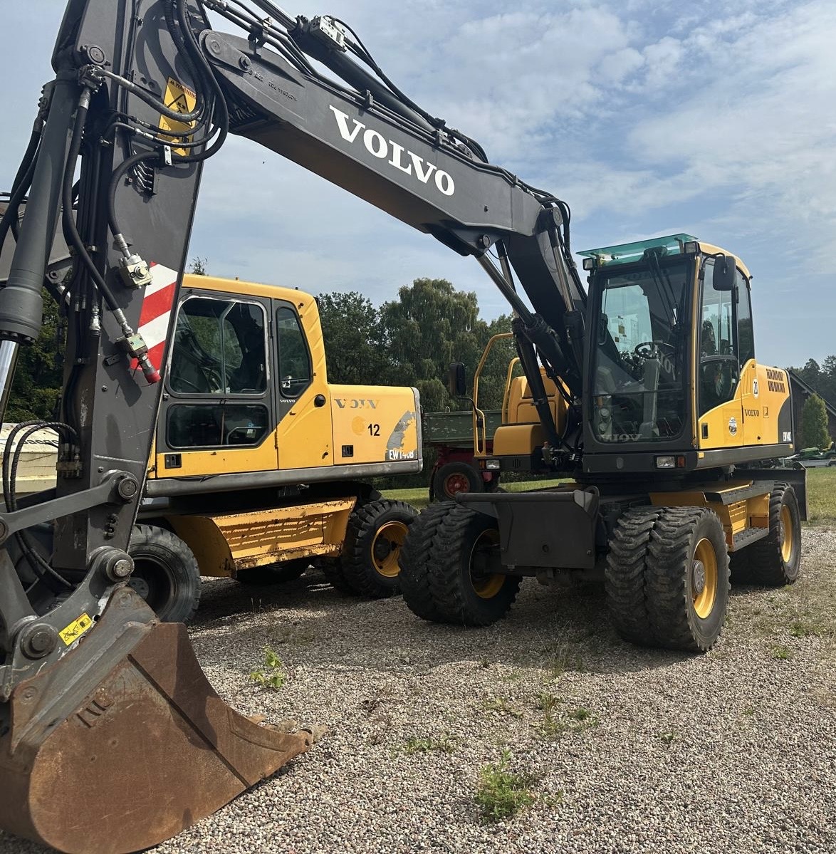 Two yellow Volvo excavators parked on gravel with green trees and partly cloudy sky behind.