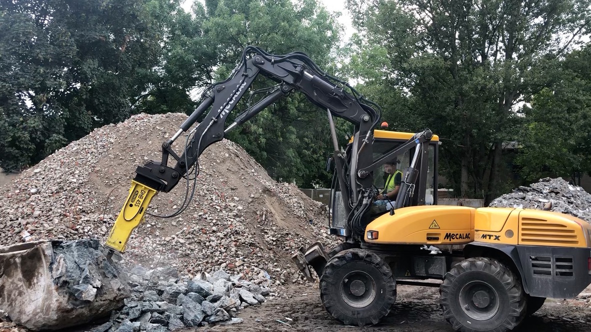 Yellow Mecalac MTX excavator breaking rocks with a hydraulic hammer attachment at a rubble site.