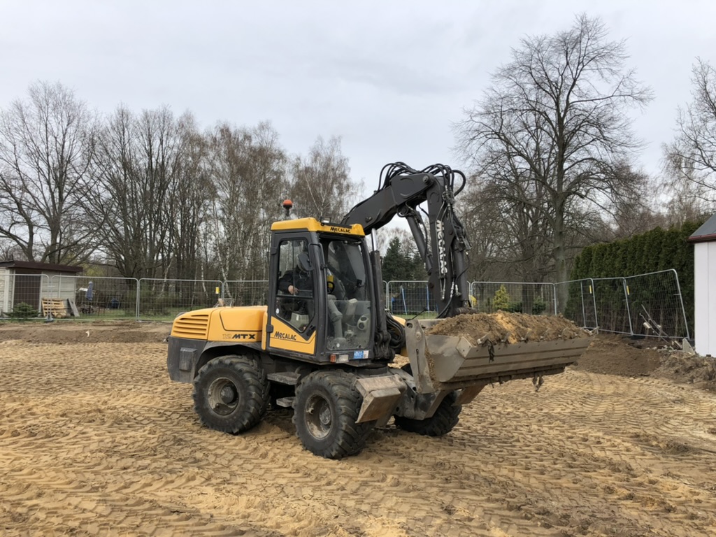 Yellow Mecalac excavator lifting a bucket full of dirt on a construction site with bare trees and fences.