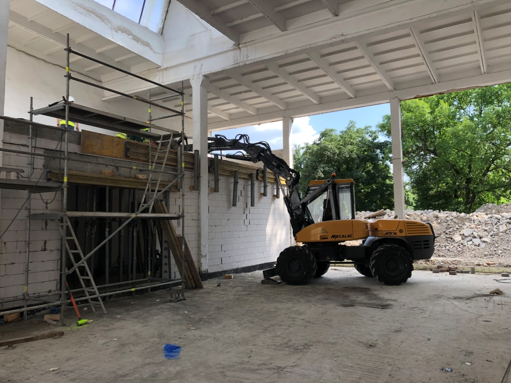 Yellow Mecalac MTX excavator inside partially constructed building near scaffolding and rubble pile.