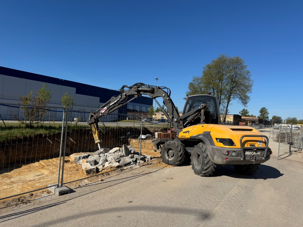 Yellow Mecalac excavator at a construction site beside a fenced trench with rubble inside.