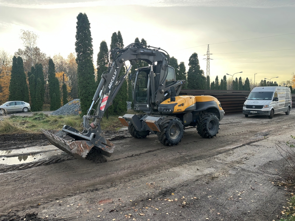 Yellow Mecalac excavator on a dirt road near a white van and tall green trees in the background.