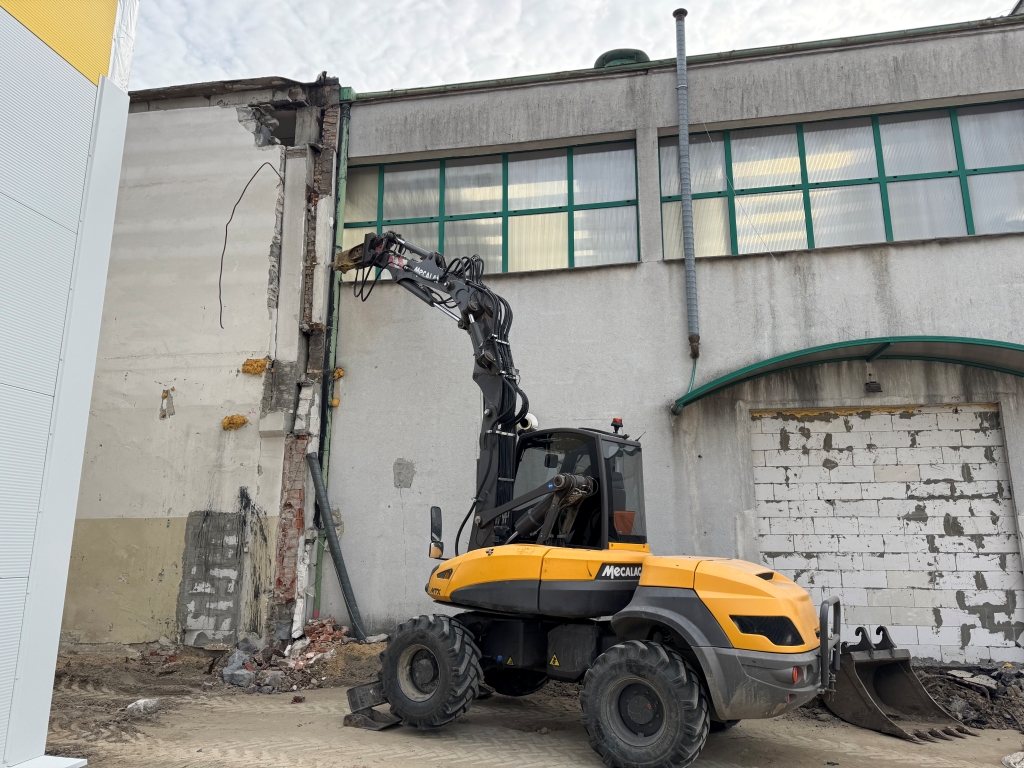 Yellow Mecalac excavator parked beside a partially demolished concrete wall with debris nearby.