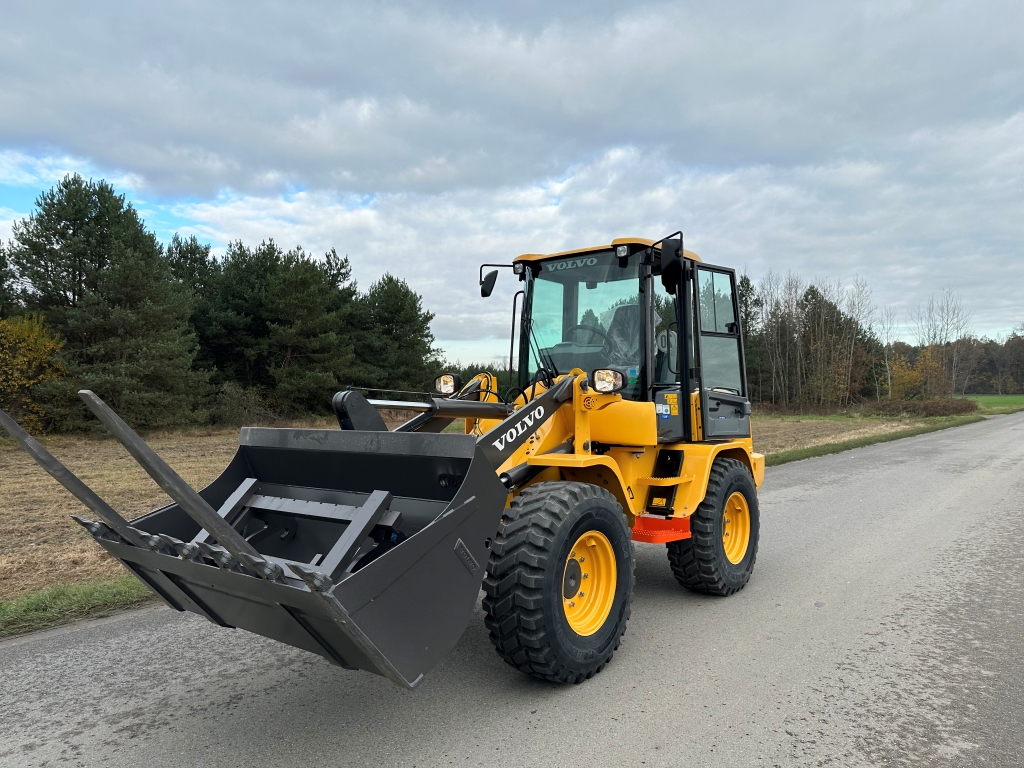 Yellow Volvo front loader with a black bucket attachment on a paved road near trees under cloudy sky.