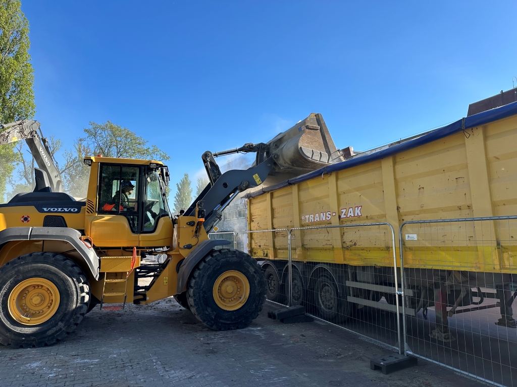 Yellow Volvo loader dumping dirt into a large yellow Trans-Zak truck behind a temporary fence under a clear blue sky.