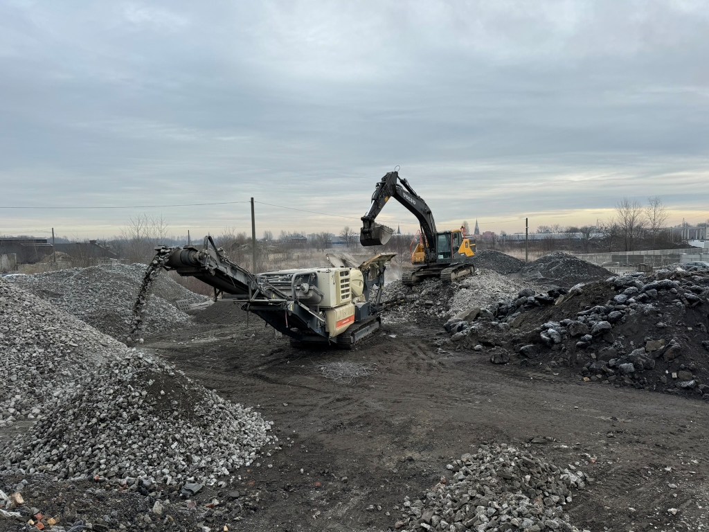 Construction site with excavator loading gravel into a machine, piles of rocks and dirt around.
